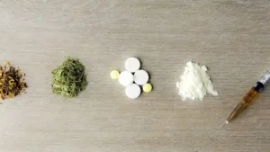 Dried leaves, green herbs, white tablets, white powder and a syringe laid in a row on a wooden surface, showing kinds of