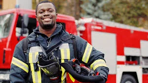 Smiling firefighter in protective gear holding a helmet, with red fire trucks parked behind him