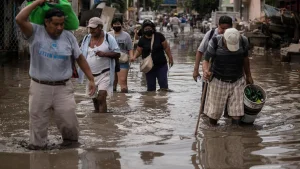 Adults wade through knee-deep floodwater along a flooded street, one carrying a bag overhead and another a bucket