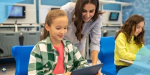 Smiling teacher leaning over a girl using a tablet in a classroom, while another girl sits nearby, showing safe school