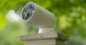 White wireless security camera mounted on a stone post outdoors, with green trees blurred behind it