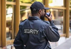 Security guard seen from behind in a cap and dark uniform with a SECURITY patch, speaking into a handheld radio