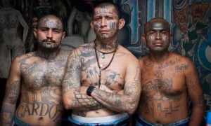 Three shirtless men with tattoos covering their faces, chests and arms stand side by side, looking at the camera.
