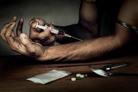 Forearm and open hand with a syringe being pushed into the arm, beside loose pills and a spoon on a dark table