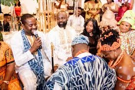 Guests in traditional attire at a ceremony, a smiling man in white and blue speaking into a microphone while others look on