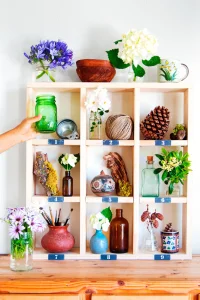 Wooden shelf of small compartments holding vases, glass jars, flowers, a pine cone and a ball of twine