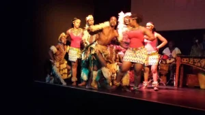 Performers in colourful traditional costumes dancing on a dark stage, with a bare-chested man at the centre