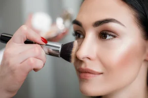 Make-up artist's hand brushing powder onto the cheek of a smiling woman with dark hair