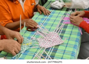 Several people weaving thin pink strips together on a table with a green checked cloth