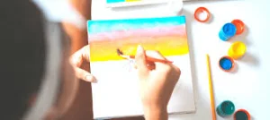 A child paints a sunset of yellow, orange and blue stripes on a small canvas, with paint pots beside it, showing visual art.