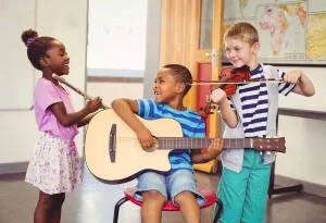 Three smiling children in a classroom, one playing a guitar, one a violin and one holding a string instrument, showing music
