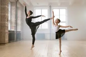 Two ballet dancers in black, a woman and a girl, balancing with raised legs in a bright studio