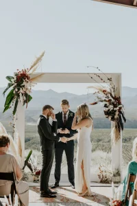 A couple holding hands facing an officiant under a floral arch outdoors at a wedding, with mountains behind