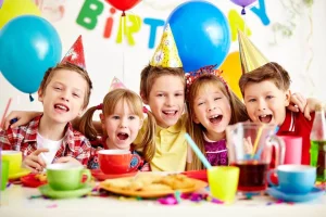 Four smiling children in party hats around a table with cake and drinks, under balloons and a birthday banner