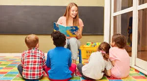 A smiling teacher reads a picture book to four young children seated on a colourful mat in a classroom