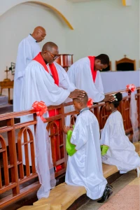 Two children in white robes kneel at a church rail while men in white robes and red stoles place hands on their heads