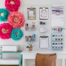Craft corner with a pegboard holding clipboards and tubs of supplies, paper flowers on the wall and a white desk