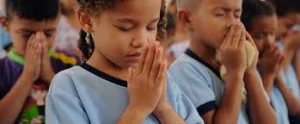 Children with eyes closed and palms pressed together in prayer, a young girl in the front