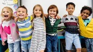 Six smiling young children standing in a row with their arms around each other's shoulders in a classroom